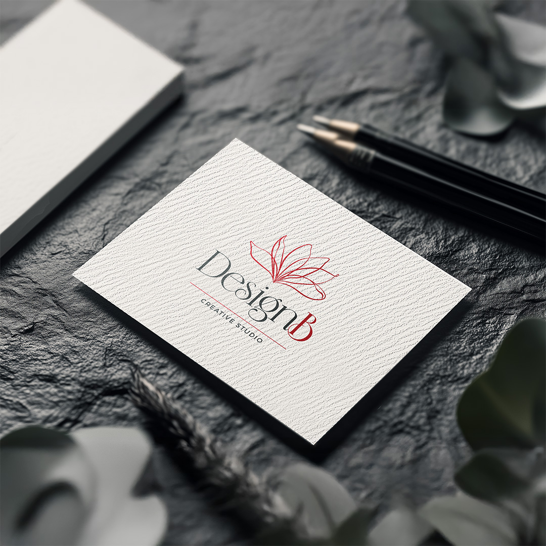 Elegant DesignB Creative Studio logo with red floral line illustration above stylised typography on textured white business card mockup, surrounded by pens and eucalyptus leaves on dark slate background